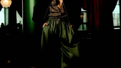 Portrait de mode d’une femme en tenue élégante aux tons sombres, debout dans un intérieur raffiné éclairé par des luminaires décoratifs, avec une esthétique cinématographique et un clair-obscur marqué.