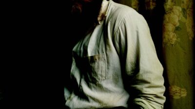 Portrait artistique d’un homme barbu portant une chemise claire, photographié dans une ambiance sombre avec éclairage dramatique, inspirée des codes du portrait éditorial et de la photographie de mode.