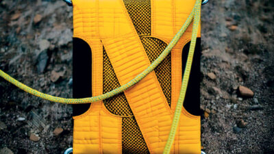 Photographie publicitaire d’un accessoire textile jaune inspiré de l’équipement d’escalade, entouré de cordes et de mousquetons sur un terrain rocheux, illustrant la robustesse et l’univers de l’outdoor.