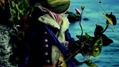 Illustration fantastique d’un personnage à tête de lézard vêtu d’un costume d’inspiration aristocratique et d’un chapeau fleuri, tenant une plante au bord de la mer dans une ambiance poétique et surréaliste.