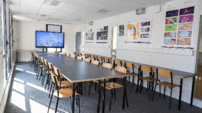 Grande salle de cours lumineuse avec longues tables, écran interactif et affiches de projets étudiants