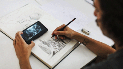 Étudiant réalisant un croquis au crayon dans un carnet de dessin avec référence photo sur smartphone