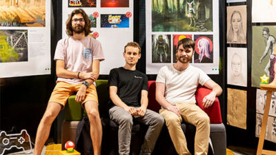 Portrait de trois étudiants devant une exposition de projets artistiques et jeux vidéo
