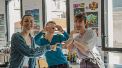 Trois étudiantes souriantes posant dans une salle d’exposition de projets étudiants