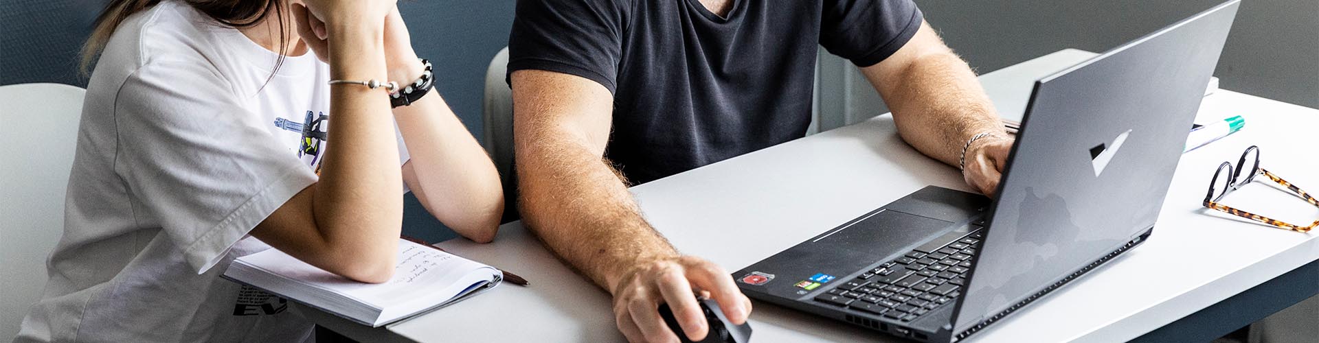 Deux personnes travaillant ensemble sur un ordinateur portable noir posé sur un bureau blanc avec un carnet de notes.