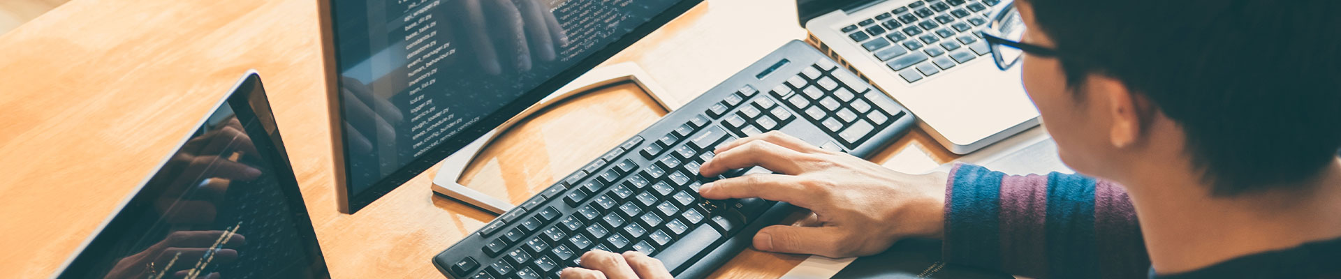 Jeune qui tape du code sur un clavier, devant plusieurs écran