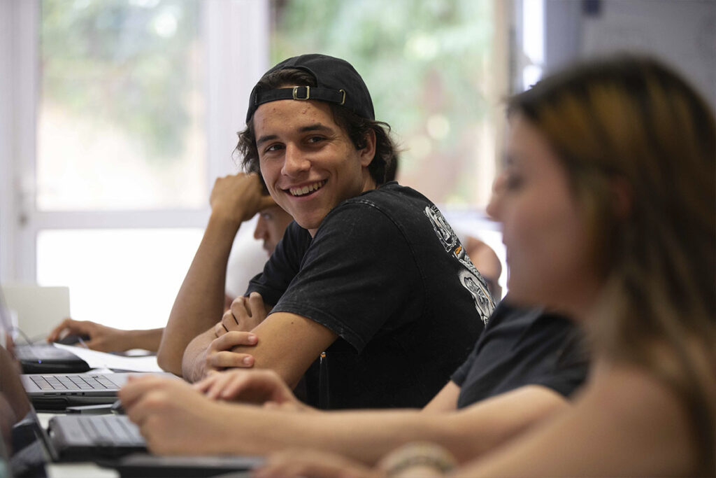 Un jeune étudiant avec une casquette à l'envers souriant en classe pendant un cours sur ordinateur.